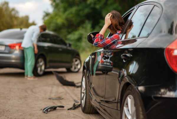 Two drivers standing beside damaged vehicles after a car accident on a roadway, discussing the collision while assessing visible front-end damage.