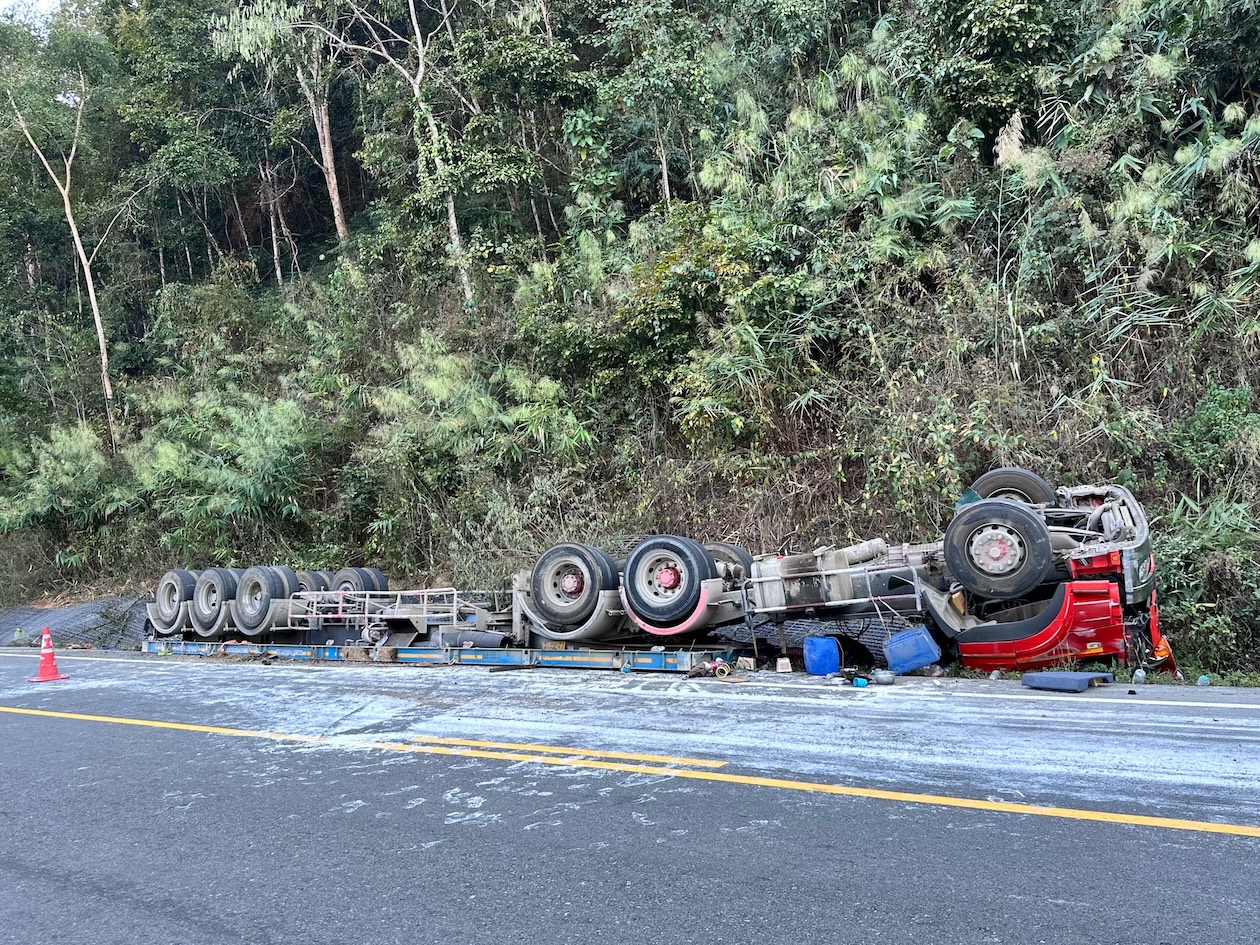 A large red semi-trailer truck lies overturned along the side of a winding mountain road after a serious rollover accident. The damaged tractor-trailer blocks part of the roadway as emergency response appears to be underway in a rugged landscape.