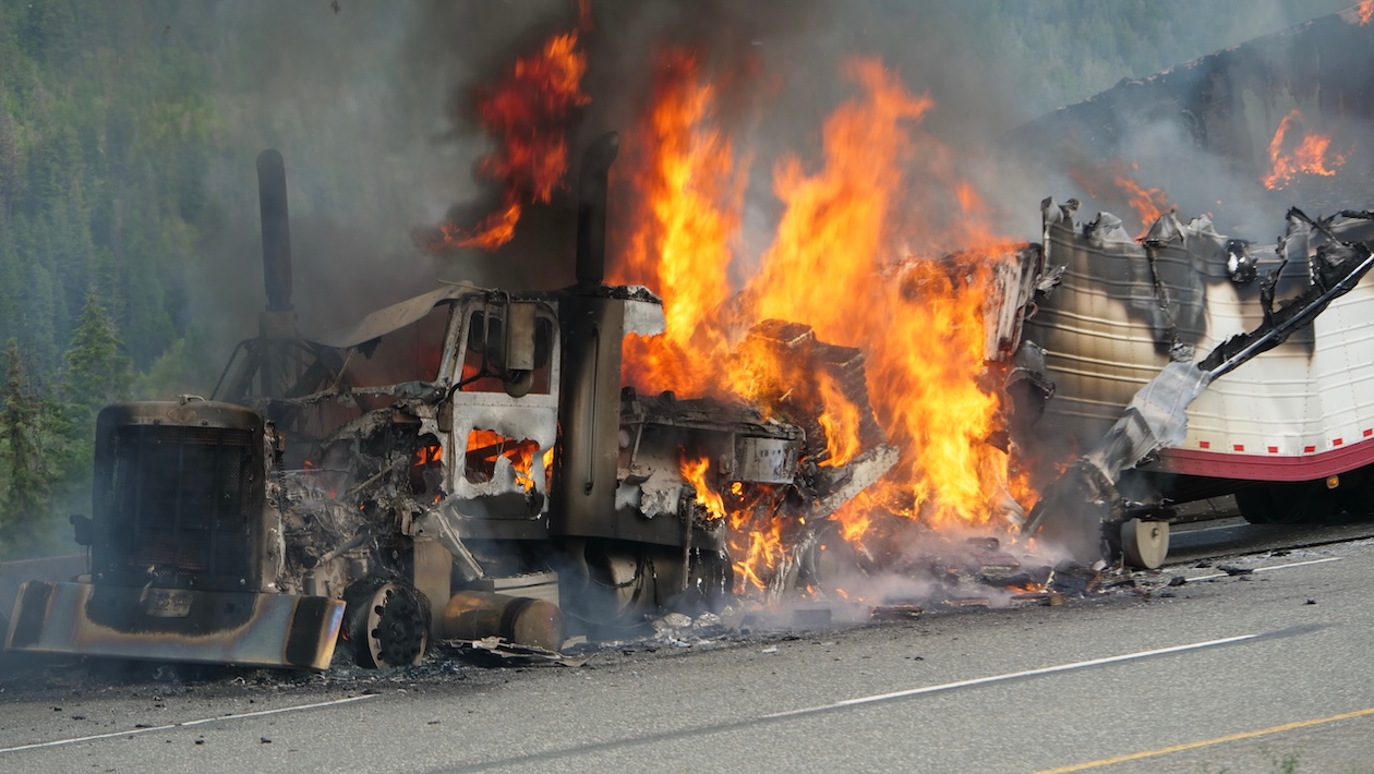 A semi-truck is engulfed in flames on the side of a highway as thick black smoke rises into the sky. Emergency responders are at the scene while traffic is affected by the intense vehicle fire.