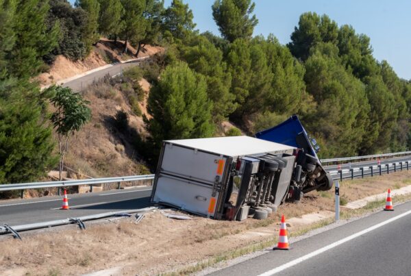 An overturned refrigerated semi-trailer truck lies on its side in the highway median near an exit ramp after a serious accident. Emergency response vehicles are on scene as traffic continues on the adjacent roadway under clear skies.