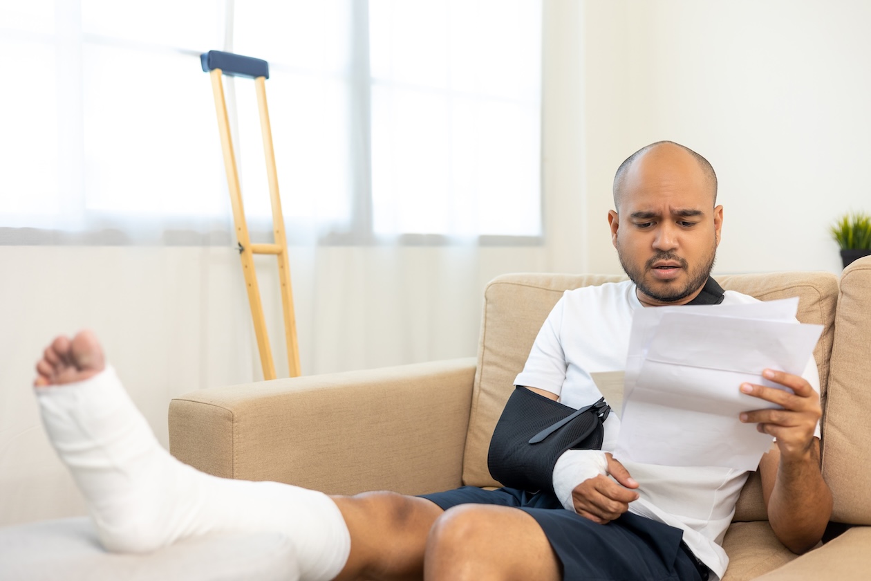 Injured man with a broken arm and leg reviewing paperwork while recovering at home after a car accident.